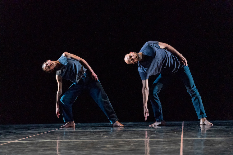 two dancers wearing blue pants and tshirts lean to one to the right with their right arm extending toward the floor as they stare out at us. Their left arm lies close to their body and the place their left hand on their hip.
